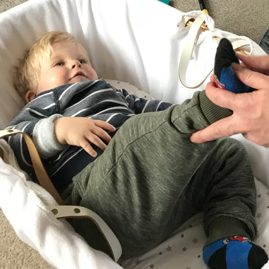 blonde haired white toddler in Baby Moses basket receiving reflexology by adult caregiver.